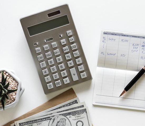A top-down view image of a calculator on a white table. To the right of the calculator is a notepad with a pen on top of it. To the left of the calculator, there is a small succulent in a pot. Below the calculator is a stack of US one-hundred dollar bills.